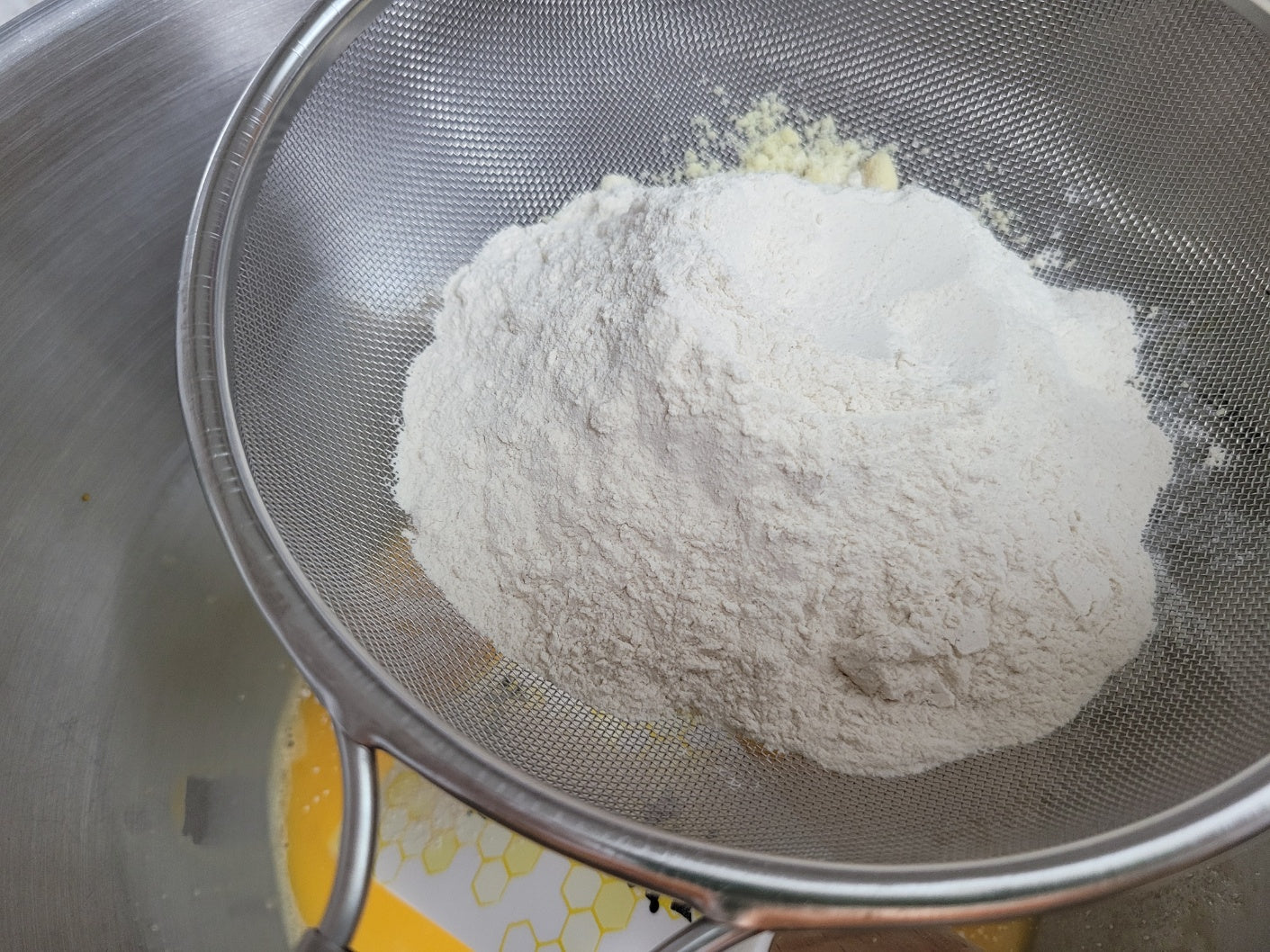 Flour being sifted through a metal sieve into a bowl.